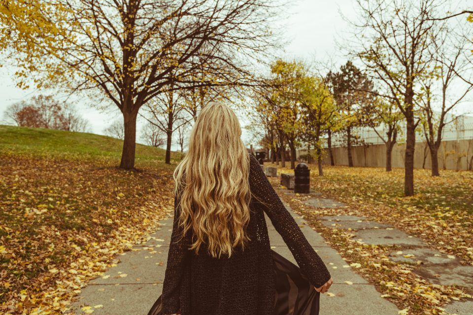 Femme aux longs cheveux blonds marchant de dos dans une allée de parc couverte de feuilles mortes