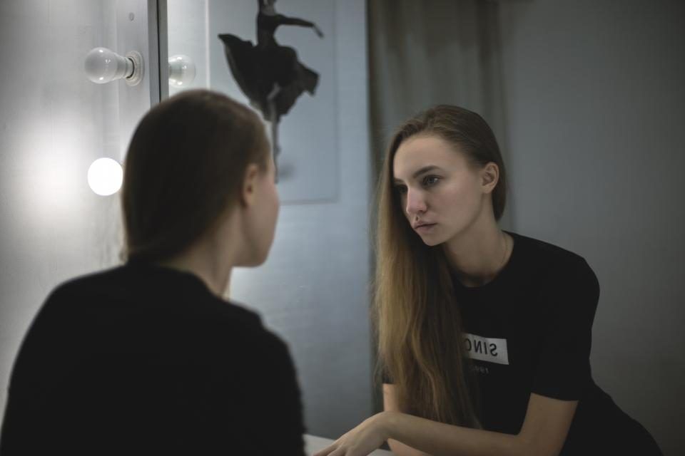 Jeune femme aux longs cheveux châtains observant son reflet dans un miroir de loge entouré d’ampoules